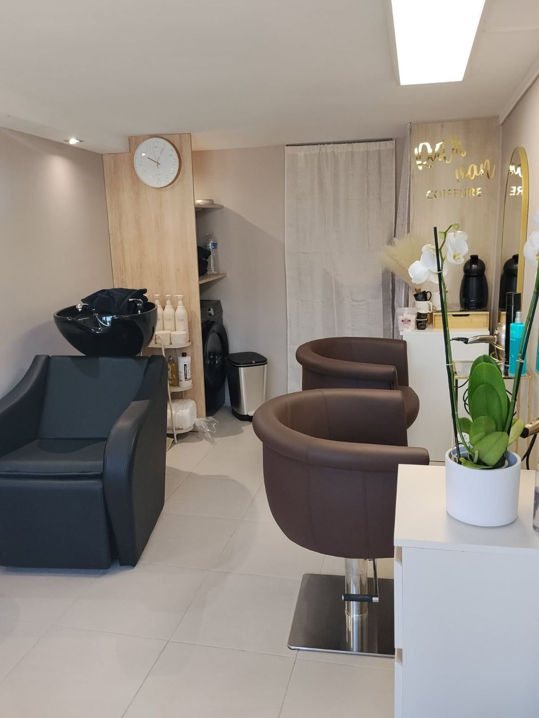 L'intérieur du salon: deux fauteuils bruns arrondis, un bac de lavage noir, un panneau de chêne clair et le lettrage doré Ruth Van Coiffure sur le mur du fond.