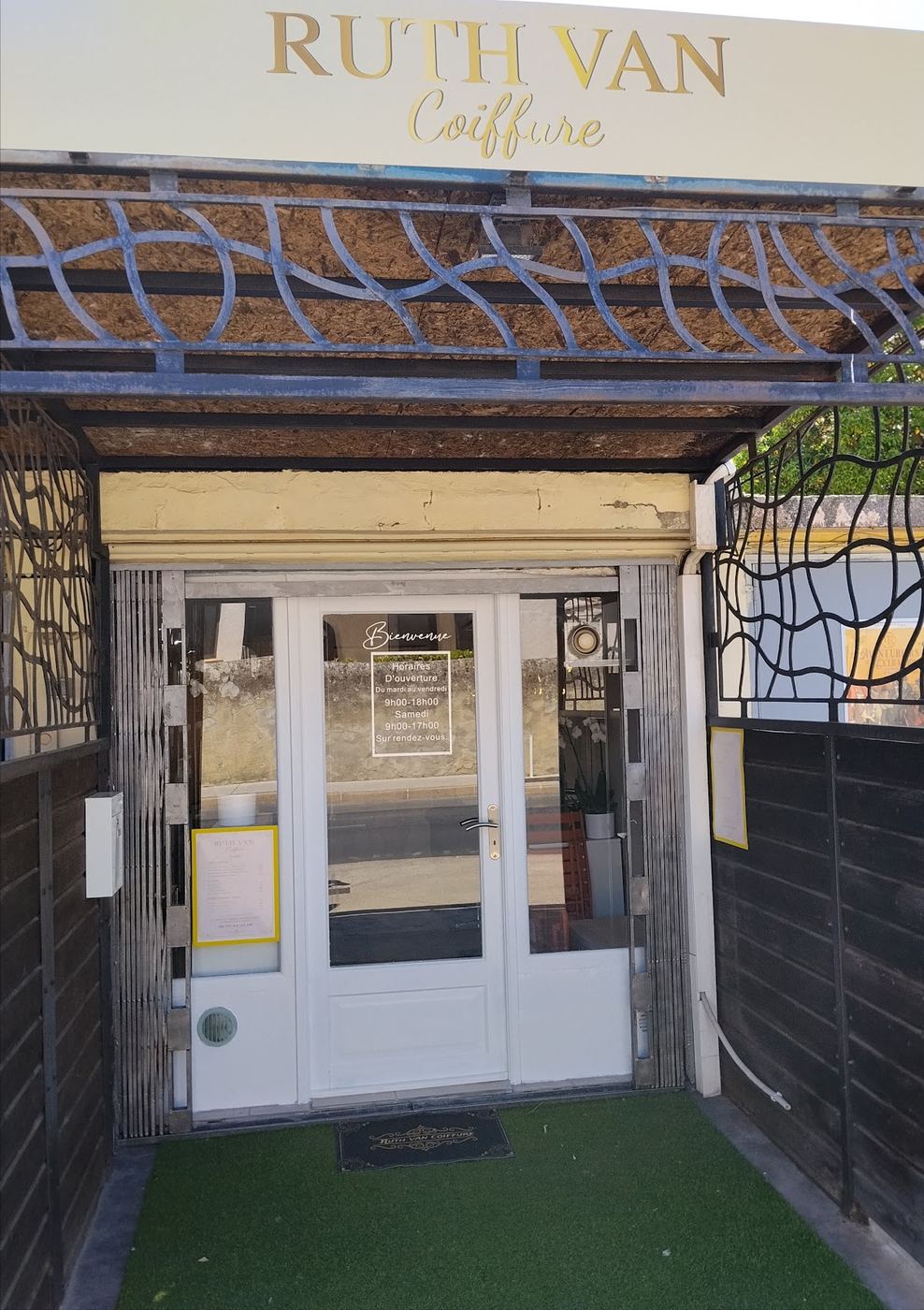 L'entrée du salon Ruth Van Coiffure sous une pergola en fer forgé noir, avec l'enseigne dorée RUTH VAN Coiffure sur un bandeau crème.